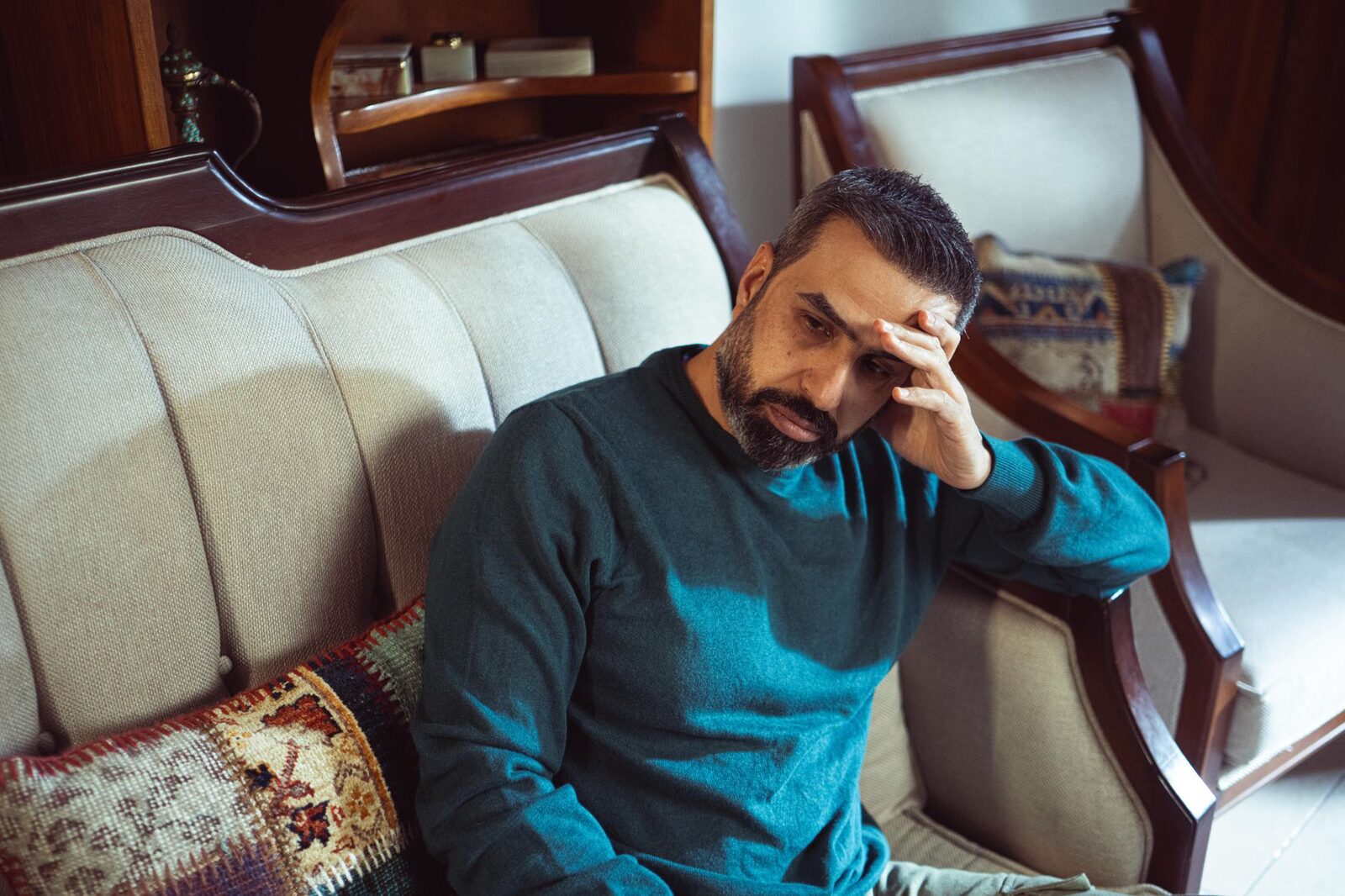 A pensive adult man in a teal sweater sits on a vintage sofa indoors, lost in thought.