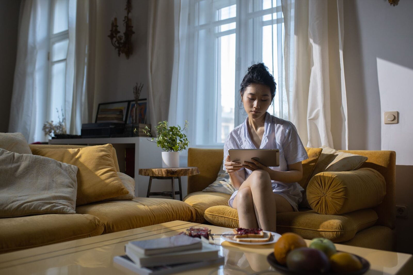 Asian woman relaxing at home, using a tablet on a cozy sofa in a well-lit living room.